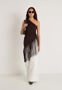 Zwarte one-shoulder top met franjes, gecombineerd met witte brede broekspijpen. Model draagt een kleine tas en donkere zonnebril.