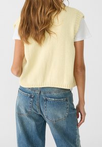 Żółty dziergany top bez rękawów z fakturowanym wzorem, w połączeniu z wysokim stanem niebieskich dżinsów z tylnymi kieszeniami i efektami przetarcia.