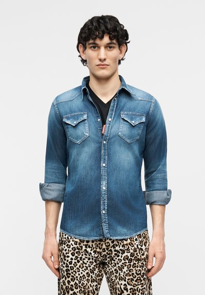 Άνδρας με σγουρά μαλλιά, φοράει τζιν πουκάμισο με γυρισμένα μανίκια πάνω από μαύρο τοπ και παντελόνι με λεοπάρ σχέδιο, στέκεται μπροστά σε απλό φόντο.