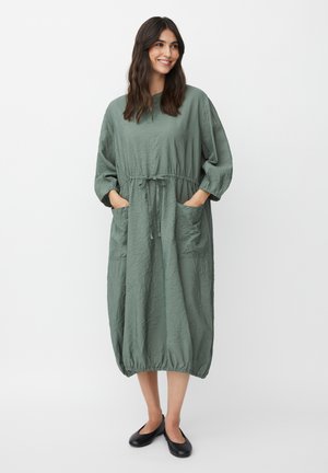 Frau steht mit den Händen in den Taschen, trägt ein lockeres, zerknittertes grünes Midi-Kleid und schwarze flache Schuhe, lächelt und schaut zur Seite.