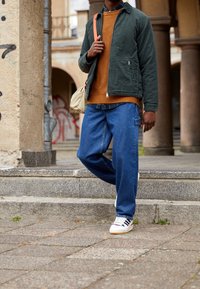 Groene jas over een oranje trui, blauwe losse jeans en witte sneakers met donkere strepen. Casual stadsoutfit met een relaxte pasvorm.