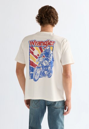 Man draagt een wit Wrangler T-shirt met een vintage-achtige afbeelding van een motorcoureur en een zonnestraalontwerp op de achterkant, gecombineerd met een blauwe spijkerbroek.