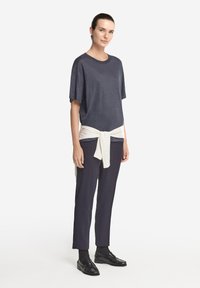 Marinblå kortärmad topp med en lös passform, tillsammans med mörka byxor; krämfärgad tröja knuten i midjan; modell som bär blanka svarta skor.