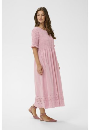 Femme portant une robe midi rose avec un haut smocké et un ourlet ajouré, associée à des ballerines roses à bride cheville, debout sur un fond blanc.