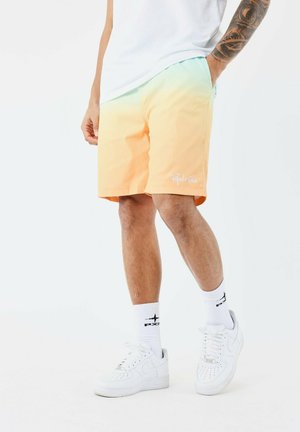 Bañador en degradado de naranja a turquesa con un logo blanco, combinado con zapatillas blancas y calcetines que presentan un detalle de diseño negro.