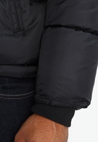 Black puffer jacket sleeve with ribbed black knit cuff, featuring a smooth, slightly shiny texture and a zipper detail on the upper arm.