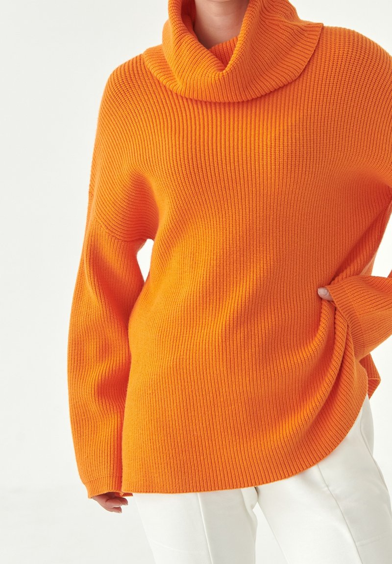 Pull à col roulé côtelé orange vif porté avec un pantalon blanc, une main glissée dans l’ourlet du pull, sur un fond clair uni.