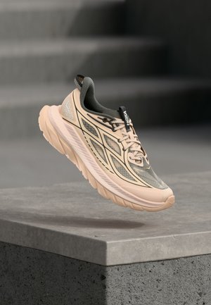 Chaussure de running rose clair et grise avec une semelle épaisse flottant au-dessus d’un rebord en béton sur un fond d’escaliers gris floutés.