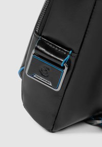 Angolo di una borsa in pelle nera con un logo in metallo argentato con accenti blu e chiusura a zip, evidenziando una texture liscia e un design elegante.