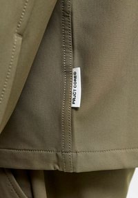 Ύφασμα ελιάς με λεία υφή, με ραμμένη ραφή και ορατή ετικέτα που αναγράφει "PROJECT CORE®".