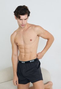Boxer shorts noirs avec une ceinture de marque, fabriqu�és en tissu léger et dotés d'un devant à boutons. Le modèle pose assis sans chemise.