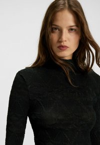 Schwarzes, strukturiertes Oberteil mit hohem Kragen, das aus einem gemusterten Stoff mit verschiedenen Oberflächendesigns und gerippten Details besteht.