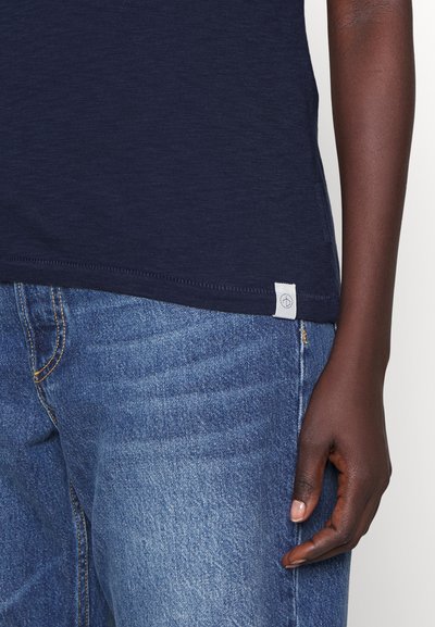 Navy blue T-shirt with a slightly textured fabric, featuring a small logo tag on the hem, paired with light blue denim jeans.