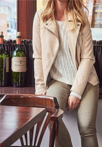 Béžová kožená motorkářská bunda, bílý pletený svetr, olivově zelené kalhoty. Dřevěný stůl a tmavý závěs v pozadí s lahvemi vína.