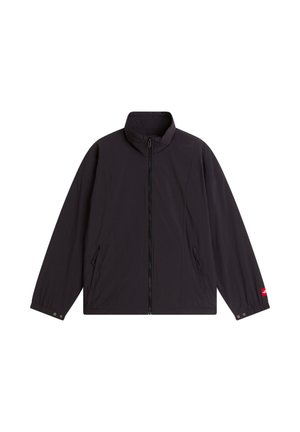 Giovane uomo con berretto giallo, giacca nera con zip, camicia scura e pantaloni grigio chiaro, in piedi su sfondo bianco.