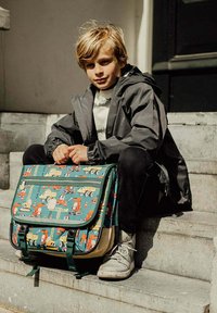 Groene boodschappentas met patronen van bouwvoertuigen, voorzien van een voorflap, dubbele ritssluitingen en verstelbare schouderbanden, geplaatst op stenen trappen.