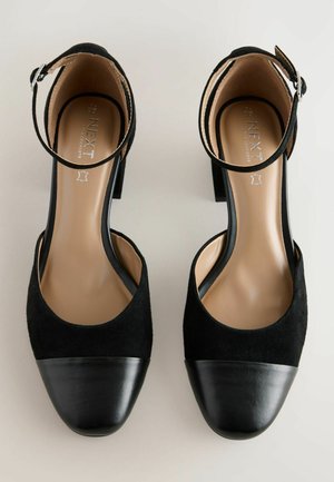 Chaussures à talons hauts noires avec une bride cheville, présentant une tige en daim et un capuchon de pied noir brillant, affichées de dessus sur un fond clair.