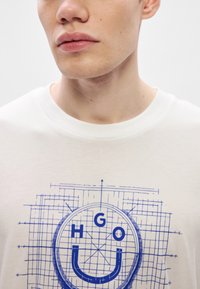 Camiseta blanca de algodón con un gráfico circular con líneas de estilo plano azul y el texto "HGO" sobre un icono de cara sonriente.