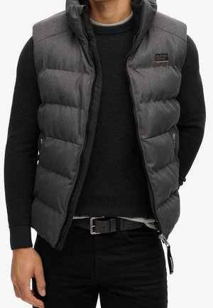 Mand iført en grå vatteret vest over en sort striktrøje og sorte bukser, hænder afslappet ved siden, Superdry-logo på vestens bryst.