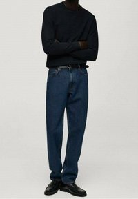 Mężczyzna stojący z założonymi skrzyżowanymi ramionami, ubrany w czarną koszulę z długim rękawem, niebieskie dżinsy z czarnym paskiem oraz czarne skórzane buty.