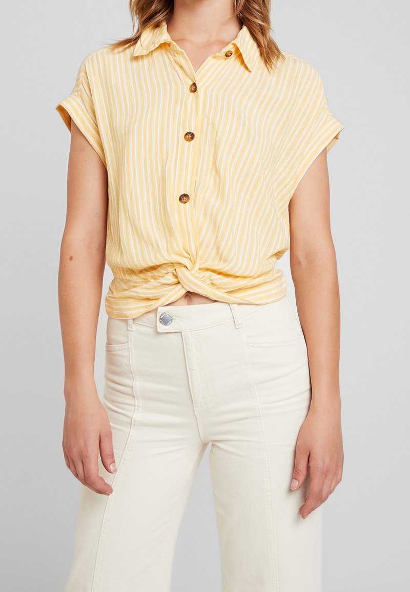 Chemise à manches courtes rayée jaune avec un devant noué, dotée d'un col et de boutons marron, associée à un jean blanc taille haute.