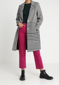 Schwarz-weißer Hahnentritt-Mantel mit einer längeren Silhouette und zwei Knöpfen, kombiniert mit knalligen pinken, cropped Hosen und schwarzen Stiefeln.