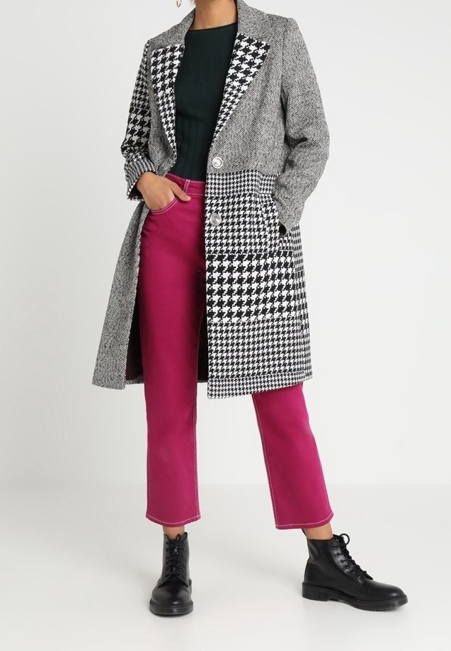 Schwarz-weißer Hahnentritt-Mantel mit einer längeren Silhouette und zwei Knöpfen, kombiniert mit knalligen pinken, cropped Hosen und schwarzen Stiefeln.