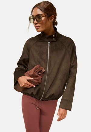 Kvinde iført en oversized brun lynlåstrøje og solbriller holder en foldet brun ruskindstaske mod en ensfarvet baggrund.
