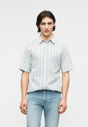 Νεαρός με σκοτεινά μαλλιά που φοράει ένα ελαφρώς γαλάζιο κοντομάνικο ριγέ πουκάμισο με κουμπιά και γαλάζια τζιν, στέκεται μπροστά σε ένα απλό φόντο.