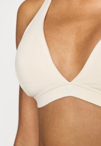 Bēšas krāsas trīsstūrveida krūšturis ar dziļu V veida izgriezumu, izgatavots no gludas auduma. Aprīkots ar platām joslām, uz kurām baltā krāsā iespiests "ICIW" logotips.