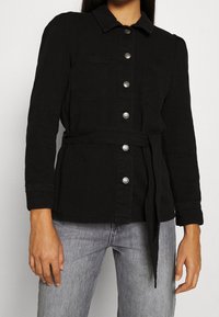 Chaqueta de mezclilla negra con mangas largas, cuello, dos bolsillos frontales y botones plateados. Incluye un cinturón atado para ajustar en la cintura.