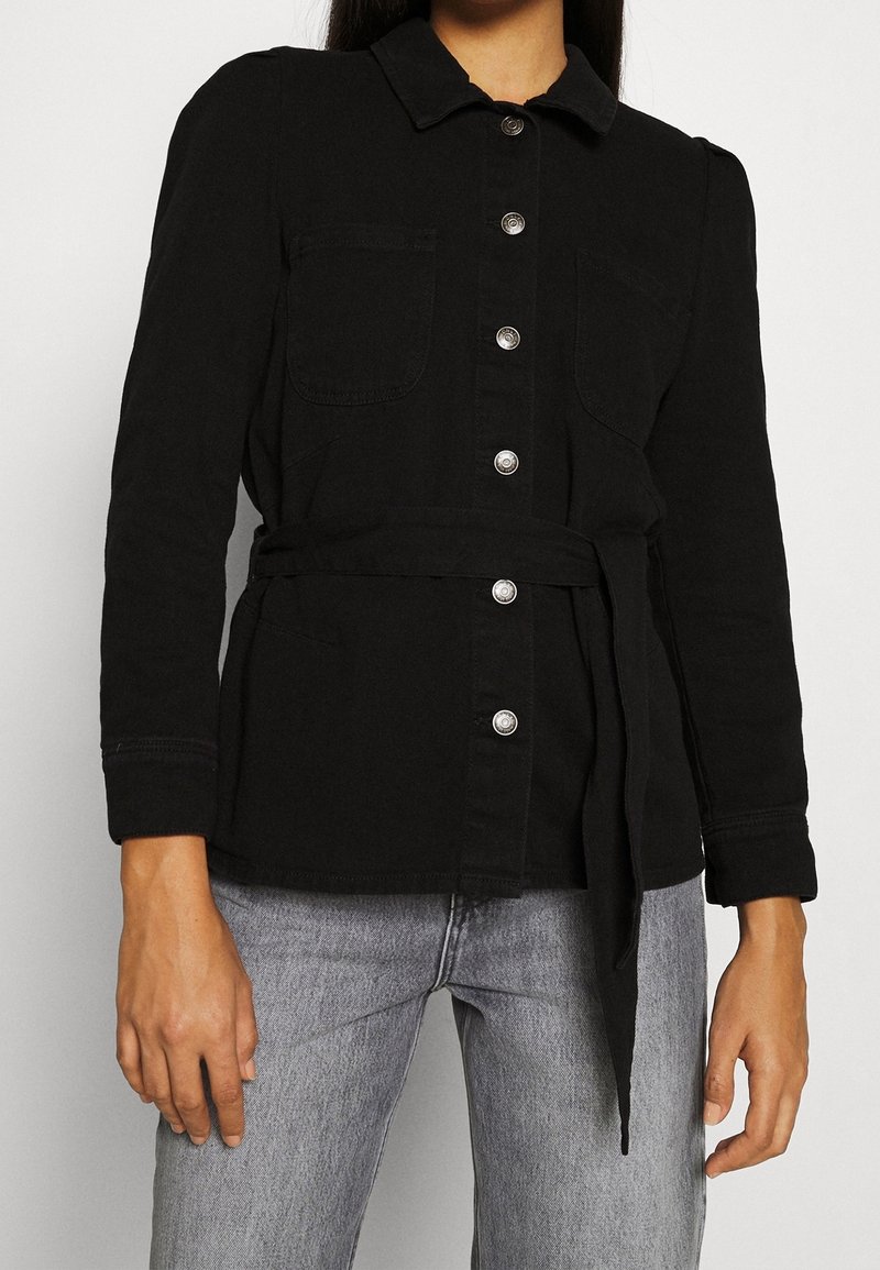 Chaqueta de mezclilla negra con mangas largas, cuello, dos bolsillos frontales y botones plateados. Incluye un cinturón atado para ajustar en la cintura.