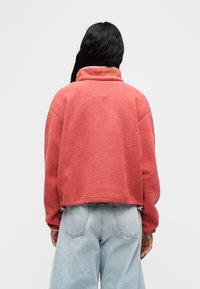 Γυναίκα με μακριά μαύρα μαλλιά που φοράει κοραλλί ροζ φλις μπουφάν και ανοιχτό μπλε τζιν παντελόνι με φαρδιά πόδια, εμφανιζόμενη από πίσω σε λευκό φόντο.