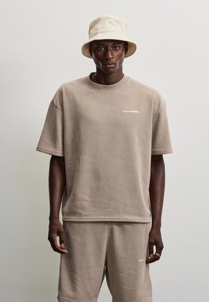 Man wearing beige ribbed short-sleeve shirt and matching shorts, with a beige bucket hat, facing forward against a plain gray background.