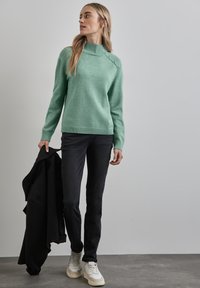 Ljusgrön stickad tröja med hög krage och knappdetaljer, i kombination med svarta jeans och vita sneakers. Slät textur, avslappnad passform.