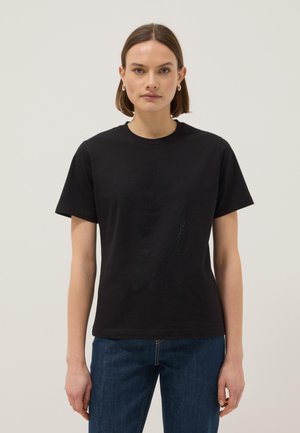 Mujer con cabello corto y castaño, que lleva pendientes de aro dorados, una camiseta negra con un diseño sutil de un oso brillante y vaqueros azules, frente a un fondo liso.