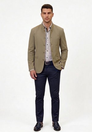 Hombre de pie con chaqueta khaki abotonada, camisa blanca con estampado, pantalones oscuros de corte slim, cinturón negro y zapatos de cuero negros, con las manos en los bolsillos.