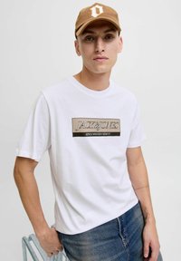 Camiseta blanca de algodón con un gráfico que incluye "JACK & JONES" y "DESDE MIL NOVECIENTOS NOVENTA". Combinada con una gorra marrón. Corte casual.
