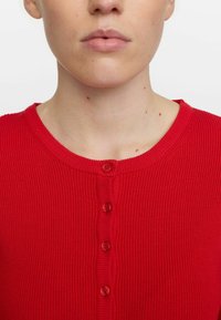 Punainen ribbikuvioinen neulecardigan, jossa on pyöreä kaula-aukko. Kardiganissa on viisi näkyvää nappia edessä ja tekstuurinen kangas.