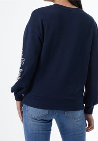 Marinblå sweatshirt med avslappnad passform, rund halsringning och puffärmar. Har pärl- och broderidetaljer på vänster ärm. Kombinerad med ljusblå jeans.