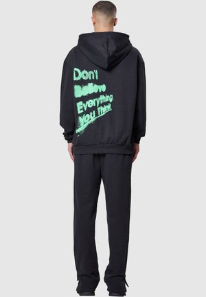 Schwarze Kapuzenjacke und passende Sweatpants aus weichem Material. Die Rückseite zeigt großen, grünen Text mit der Aufschrift "Glaube nicht alles, was du denkst."