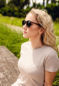 Kulaté sluneční brýle s želvovinovým vzorem a tmavými skly; texturovaná, světlá béžová triko se ribbed vzorem; trojúhelníkové zlaté náušnice; vlnité blond vlasy; zelené pozadí.