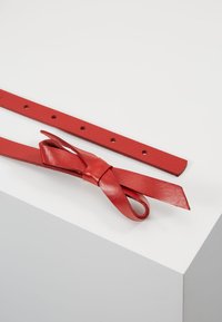 Cintura in pelle rossa con finitura testurizzata, dotata di multiple perforazioni per la regolazione e un accento decorativo a forma di fiocco a un'estremità.