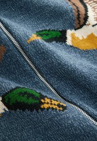 Kötött pulóver kék háttérrel, zöld, sárga és barna kacsa mintákkal, ezüst cipzár részlettel.