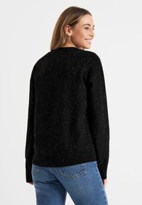 Zwarte trui met metallic spikkels, geribbelde manchetten en zoom. Heeft een ronde hals en een relaxed fit. Gepaard met blauwe spijkerbroek.