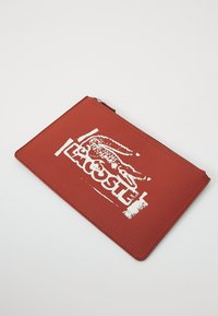 Red textured leather pouch featuring a prominent white Lacoste logo and crocodile graphic, with a zip closure at the top.