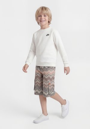 Usmievajúci sa blond chlapec kráča, má na sebe bielu mikinu Nike, vzorované krátke nohavice s vreckami a biele tenisky na jednolennom bielom pozadí.