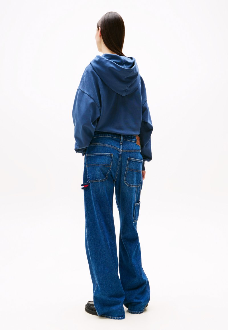 Übergroßer navy Kapuzenpullover mit Kordelzug, kombiniert mit weiten blauen Jeans, die aufgesetzte Taschen und ein logobezogenes Detail an der Seite haben.