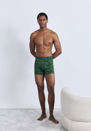 Πράσινα μποξερ με σχέδιο φύλλων, ελαστική μέση με λογότυπο "BJÖRN BORG", το μοντέλο στέκεται ξυπόλητο σε μια μινιμαλιστική σκηνή.