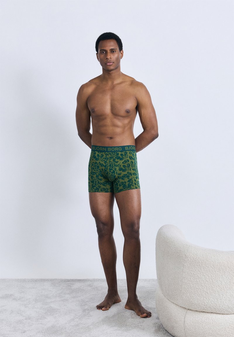 Πράσινα μποξερ με σχέδιο φύλλων, ελαστική μέση με λογότυπο "BJÖRN BORG", το μοντέλο στέκεται ξυπόλητο σε μια μινιμαλιστική σκηνή.